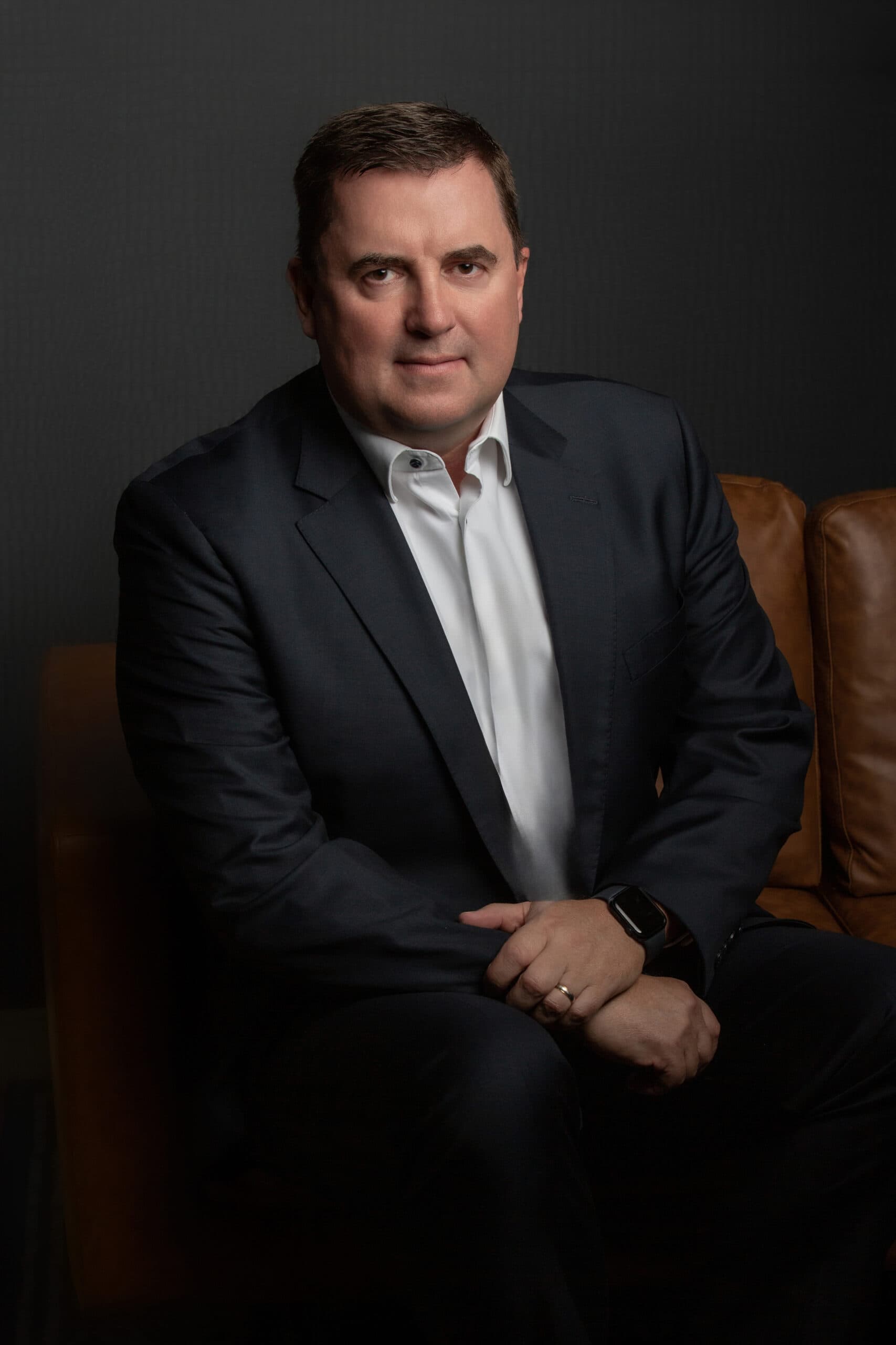 A middle-aged man in a black suit and white dress shirt sits on a chair against a dark gray background.