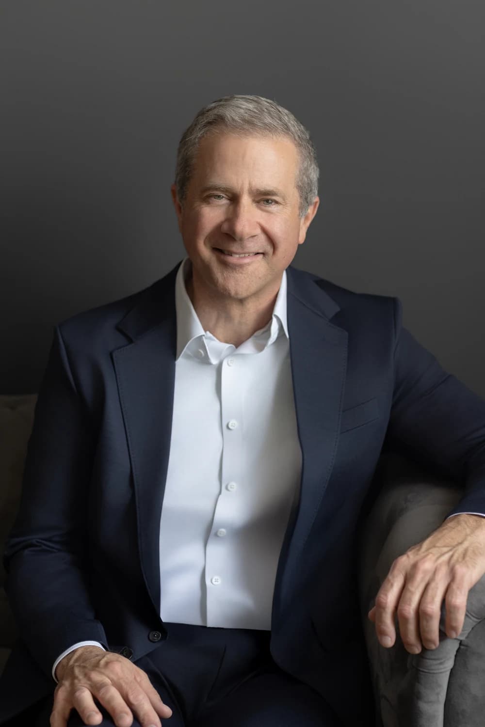 A middle-aged man in a navy suit and white dress shirt sits on a chair against a dark gray background.