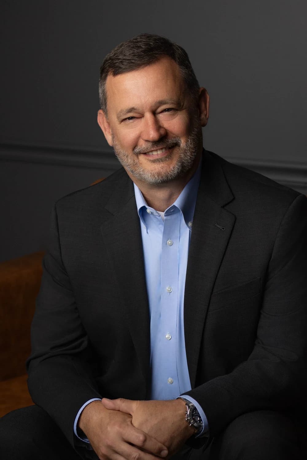 A middle-aged man in a black suit and blue dress shirt sits on a chair against a dark gray background.