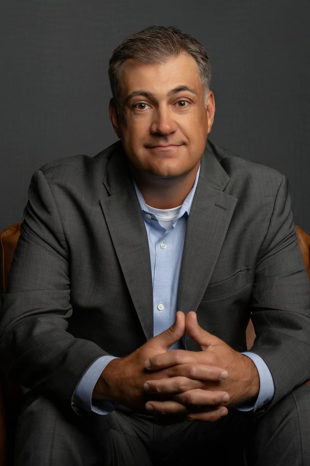 A middle-aged man in a gray suit and blue dress shirt sits on a chair against a dark gray background.